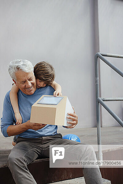 Smiling senior man holding solar house model by grandson in front of wall