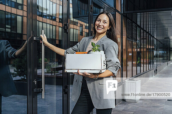 Happy businesswoman with box standing by glass door