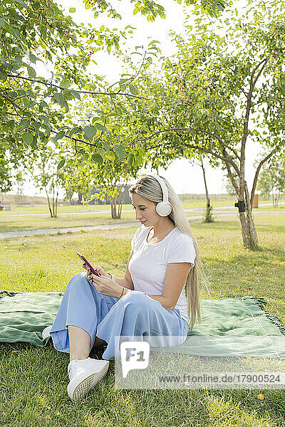 Junge Frau mit kabellosen Kopfhörern und Smartphone sitzt im Park