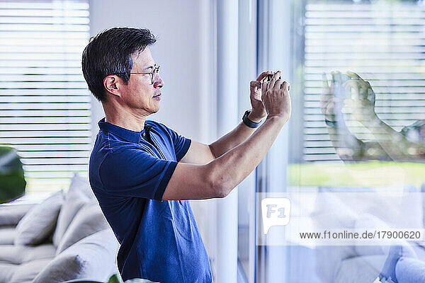 Reifer Mann fotografiert mit Smartphone durch Fenster zu Hause