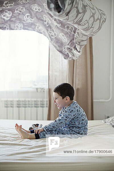 Verspielter Junge sitzt unter einer Decke in der Luft auf dem Bett