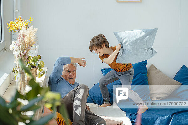 Fröhlicher Junge spielt Kissenschlacht mit Großvater auf dem heimischen Sofa