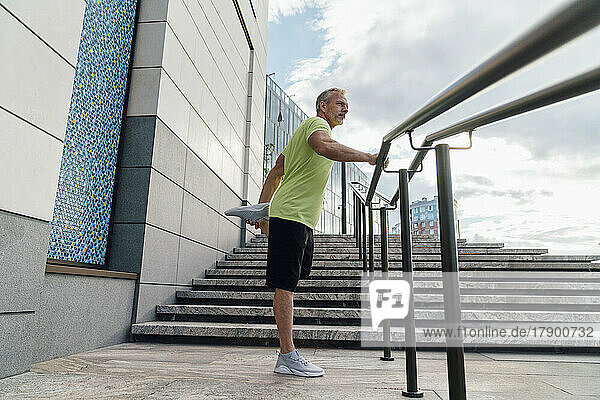 Man stretching leg standing by railing in city