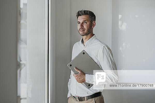 Thoughtful businessman holding laptop looking through window standing with hand in pocket at office