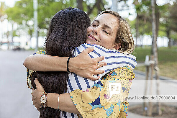 Lächelnde Frau mit geschlossenen Augen umarmt Freundin im Park