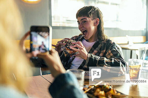 Young woman photographing girlfriend holding burger at restaurant