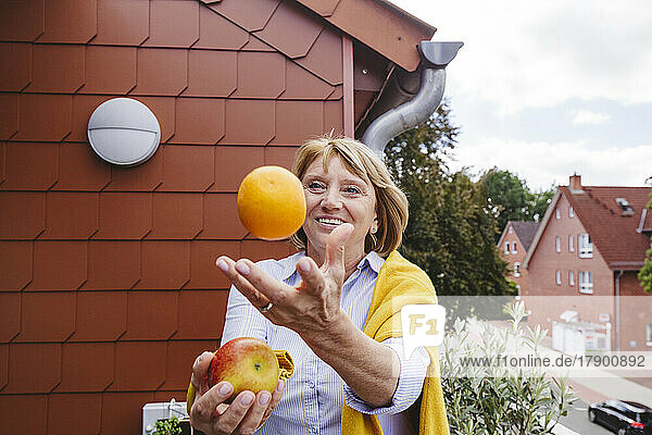 Happy senior woman juggling with orange and apple fruit in balcony