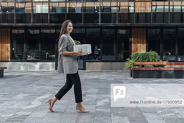 Smiling businesswoman walking with box in front of office building
