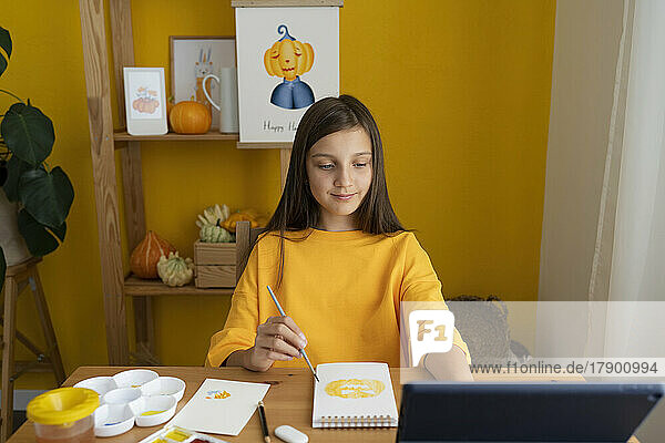Lächelndes Mädchen malt auf einem Skizzenblock und schaut sich zu Hause ein Online-Tutorial über einen Tablet-PC an