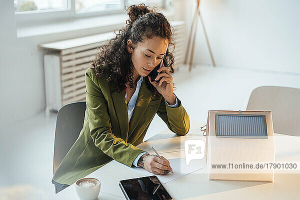 Businesswoman talking on smart phone writing at desk in office