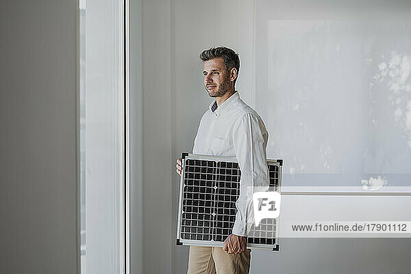 Smiling engineer holding solar panel looking through window in office
