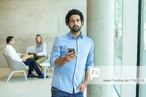 Businessman with hand in pocket holding smart phone leaning on column at office