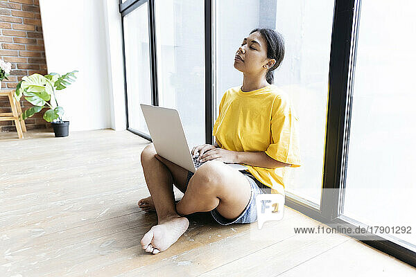 Junge Frau sitzt mit geschlossenen Augen auf dem Boden  lehnt an einem Fenster und hat einen Computer auf dem Schoß