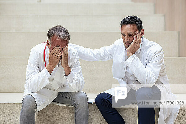 Doctor consoling upset colleague sitting on steps in hospital