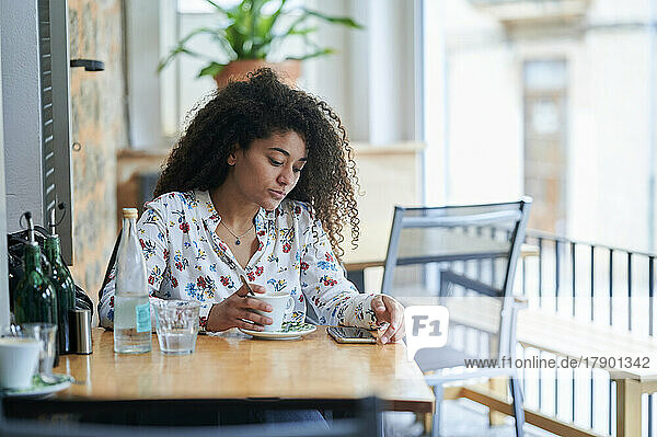 Junge Frau mit Kaffeetasse schaut im Café auf ihr Smartphone