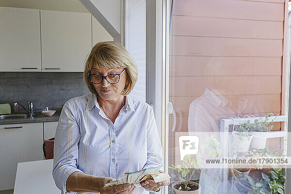 Woman counting paper currency standing by glass window at home