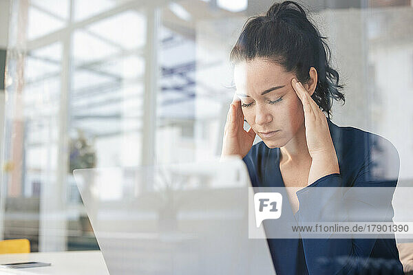 Businesswoman with eyes closed sitting in front of laptop seen through glass