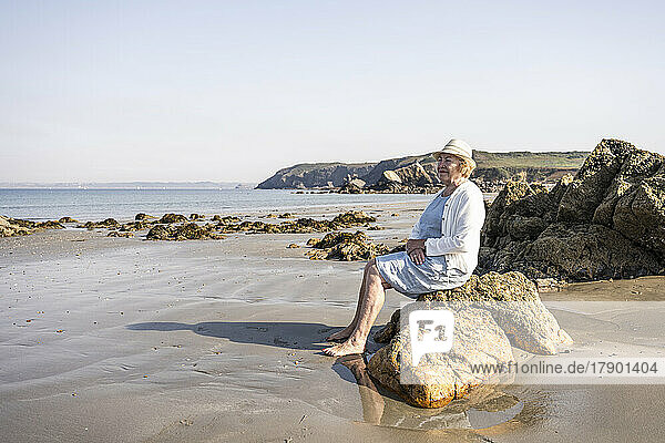 Nachdenkliche ältere Frau sitzt auf einem Felsen am Strand
