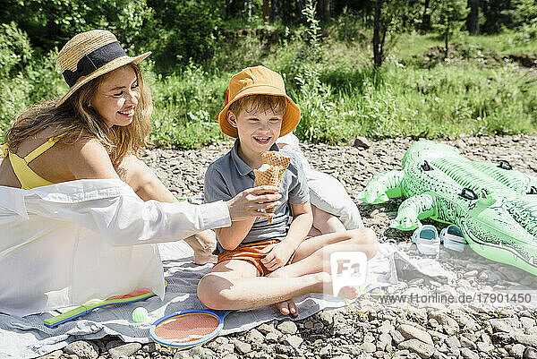 Mother giving ice cream to son sitting cross-legged at picnic on sunny day