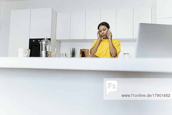 Junge Frau sitzt mit Laptop in der Küche und telefoniert
