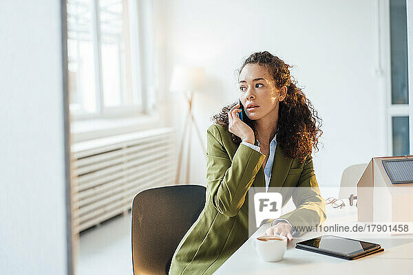 Young businesswoman talking on smart phone sitting with model home at desk in office
