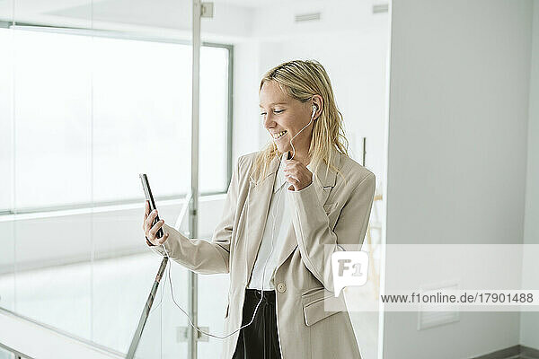 Lächelnde Geschäftsfrau blickt im Büroflur auf ihr Mobiltelefon