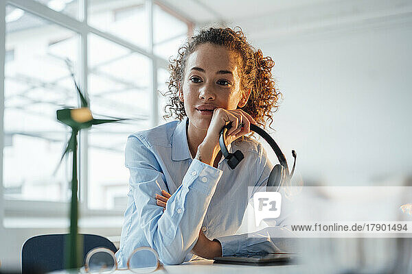 Young businesswoman holding headset looking at wind turbine in office