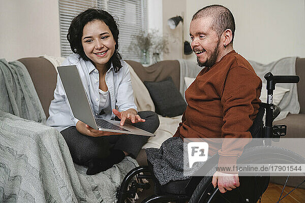 Smiling woman showing laptop to boyfriend in wheelchair at home