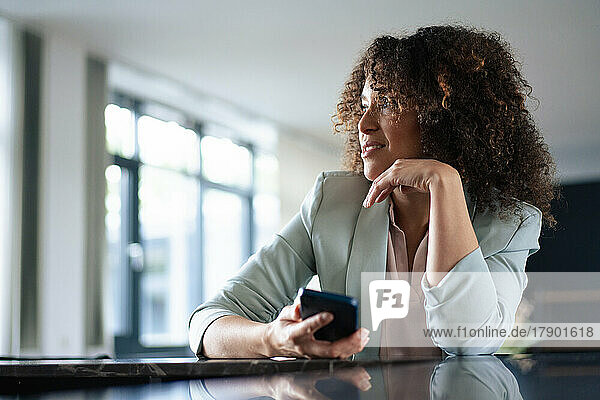 Nachdenkliche Geschäftsfrau mit Mobiltelefon am Tisch im Büro