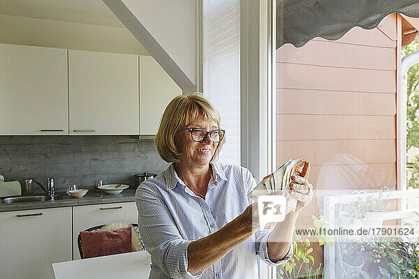 Happy senior woman counting paper currency standing at home