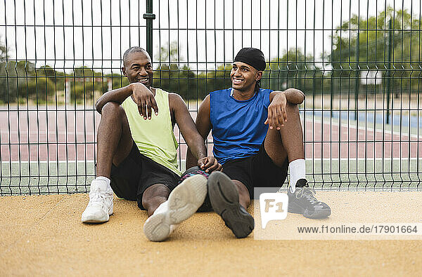 Smiling son and father leaning on fence sitting at basketball court
