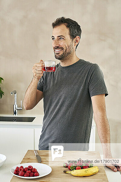 Lächelnder Mann mit Teetasse  der neben Früchten in der Küche steht