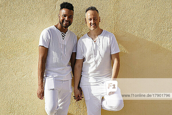 Smiling gay couple holding hands standing in front of yellow wall