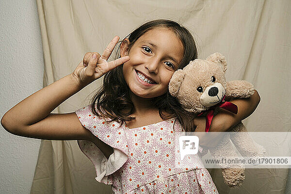 Happy girl with teddy bear gesturing peace sign