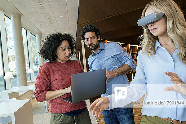 Woman wearing VR glasses by colleagues using laptop in library
