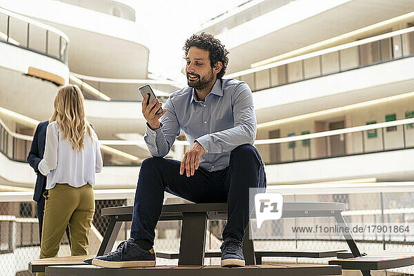Lächelnder Geschäftsmann nutzt Smartphone mit Kollegen im Hintergrund am Flur