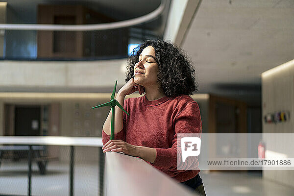 Businesswoman with eyes closed holding wind turbine on railing at office corridor
