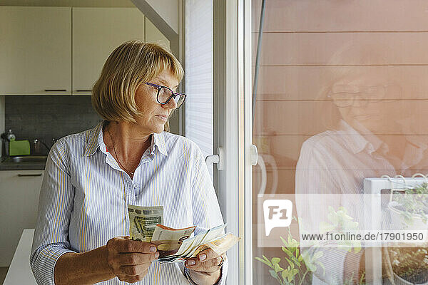 Senior woman with paper currency looking through window at home