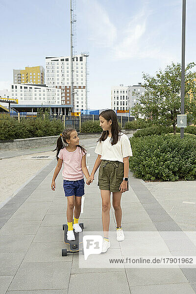 Girl holding hand of sister standing on skateboard at footpath