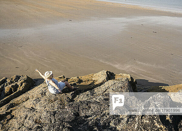 Ältere Frau mit ausgestreckten Armen sitzt auf einem Felsen am Strand