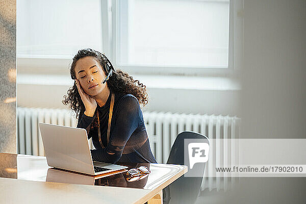 Junge Geschäftsfrau mit Headset sitzt mit geschlossenen Augen am Schreibtisch im Büro