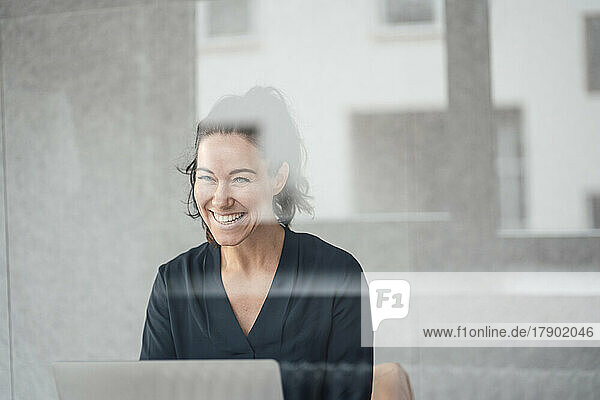 Happy businesswoman with laptop in front of wall seen through glass