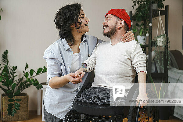 Lächelnder Mann im Rollstuhl hält zu Hause die Hand seiner Freundin