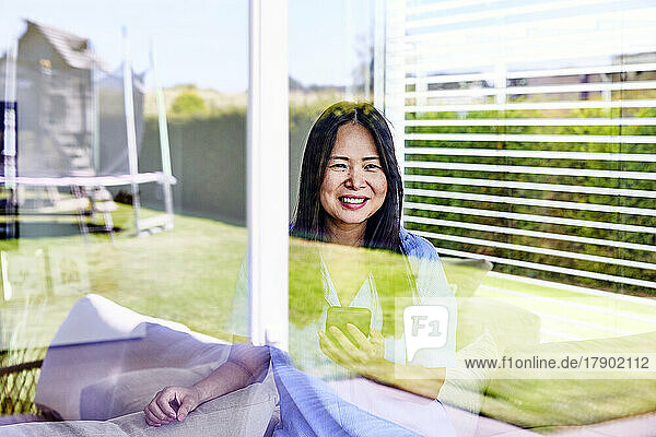 Smiling mature woman with smart phone seen through glass