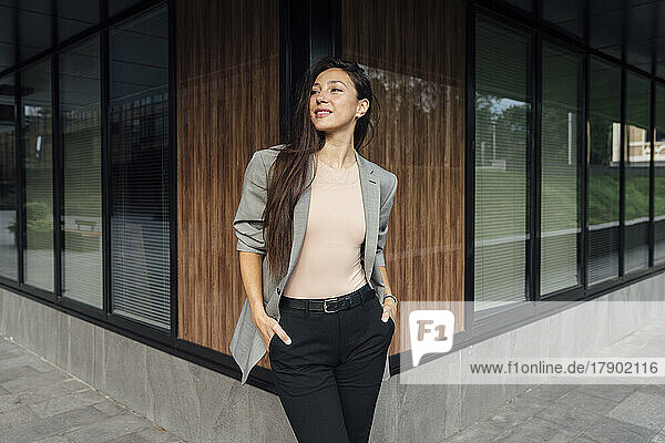 Smiling businesswoman standing with hands in pockets outside office building
