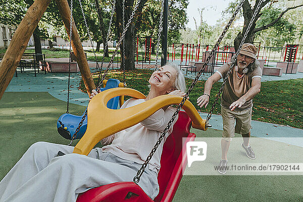 Senior man pushing woman enjoying swing at park