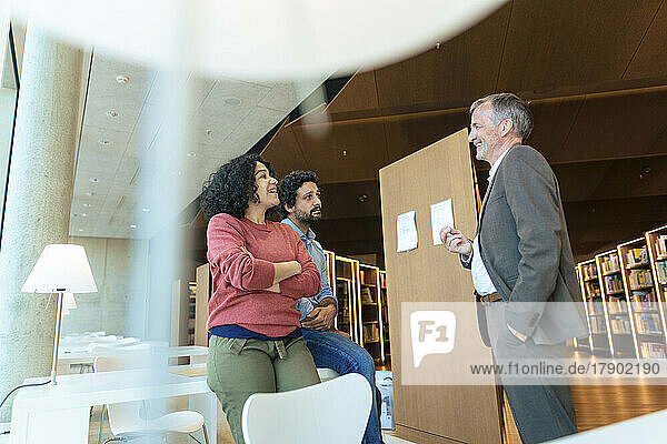 Smiling man with hand in pocket talking with colleagues in university library