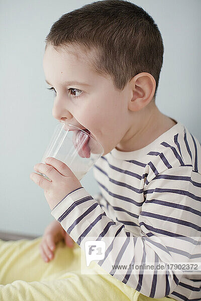 Cute boy licking glass of milk sitting by wall