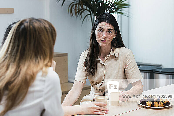 Young businesswoman discussing with colleague sitting in cafeteria