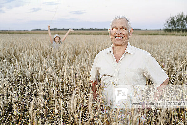 Lächelnder Bauer inmitten von Feldfrüchten und Enkelin  die sich im Hintergrund auf dem Bauernhof amüsiert